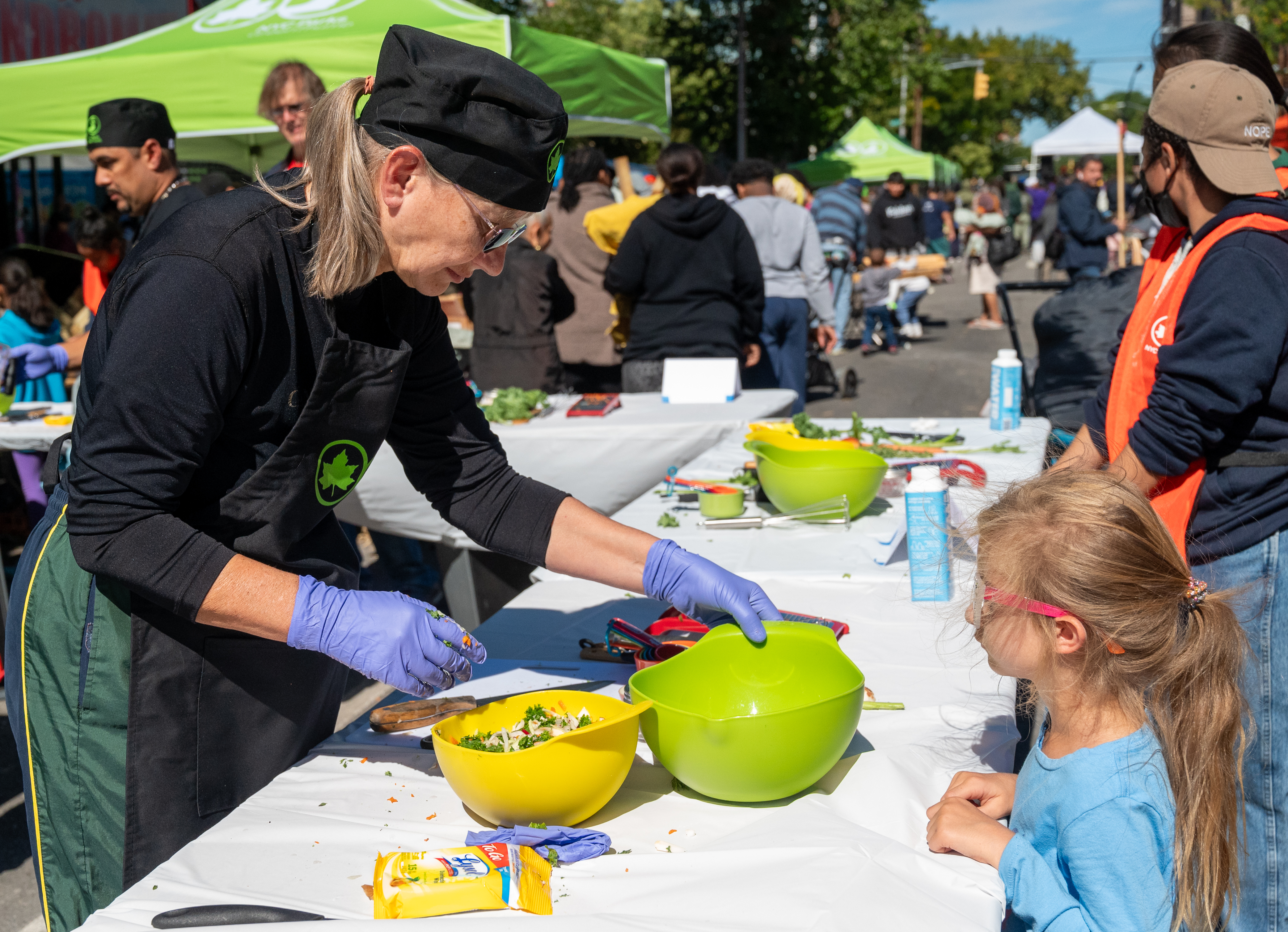 A woman mixing food in a yellow and green bowl at a long table with a white tablecloth on a city street. There is a small child leaning on the table and watching the woman cook.