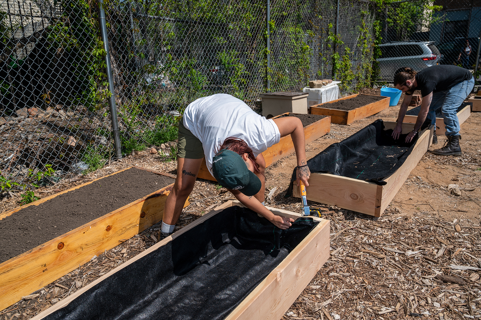 Two people are bent over raised beds in a community garden, while they fix fabric to the beds with their hands or a hammer. 