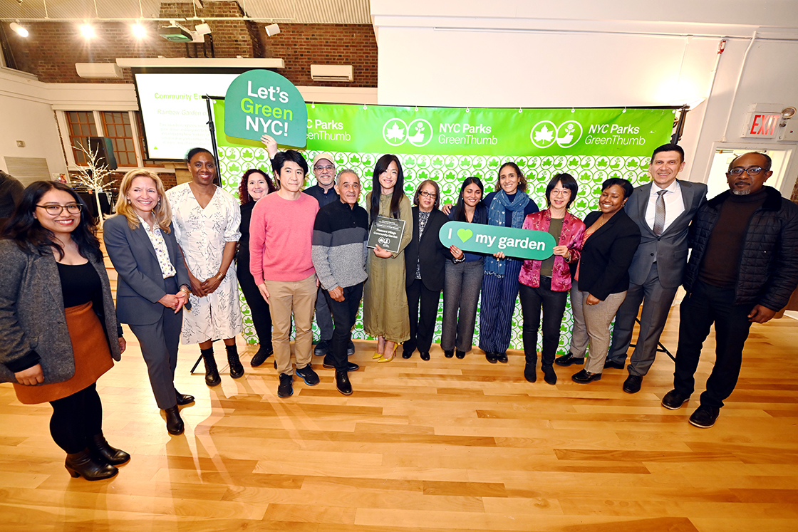 NYC Parks staff and honorees of the GreenThumb Landscape Design Award are standing in front of a bright green and white NYC Parks GreenThumb step and repeat at the 2023 Garden Recognition Awards ceremony. A person in the center is holding the award, a hunter green plaque, and one person is holding a sign saying I heart my garden.