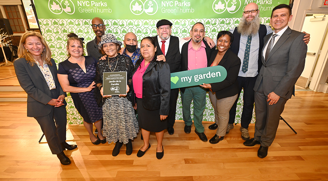 NYC Parks staff and honorees of the GreenThumb Urban Agriculture Award are standing in front of a bright green and white NYC Parks GreenThumb step and repeat at the 2023 Garden Recognition Awards ceremony. A person in the center is holding the award, a hunter green plaque, and one person is holding a sign saying I heart my garden.
