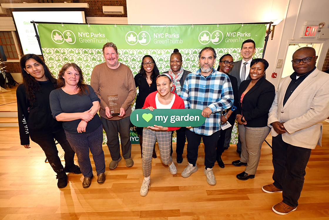 NYC Parks staff and honorees of the GreenThumb Going the Extra Mile Award are standing in front of a bright green and white NYC Parks GreenThumb step and repeat at the 2023 Garden Recognition Awards ceremony. A person is holding the glass award and one person is holding a sign saying I heart my garden.