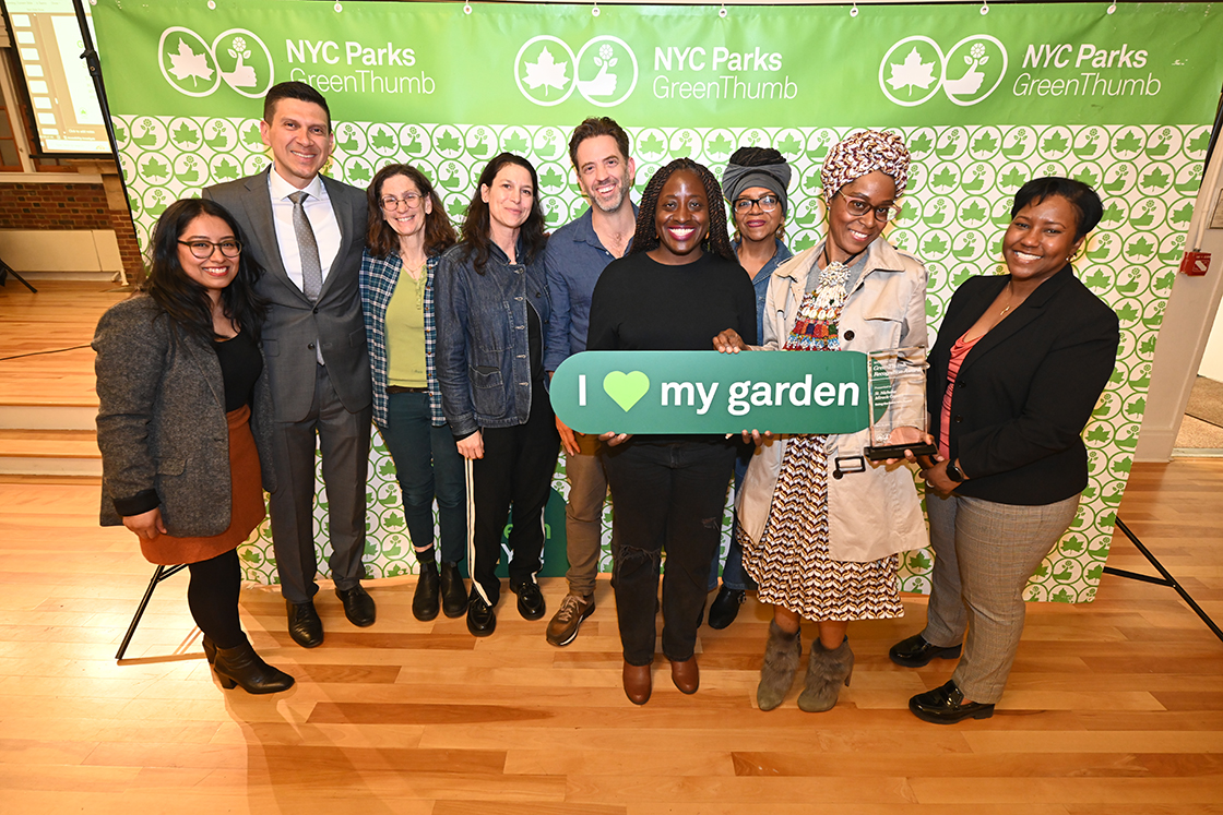 NYC Parks staff and honorees of the GreenThumb Going the Extra Mile Award are standing in front of a bright green and white NYC Parks GreenThumb step and repeat at the 2023 Garden Recognition Awards ceremony. A person is holding the glass award and one person is holding a sign saying I heart my garden.