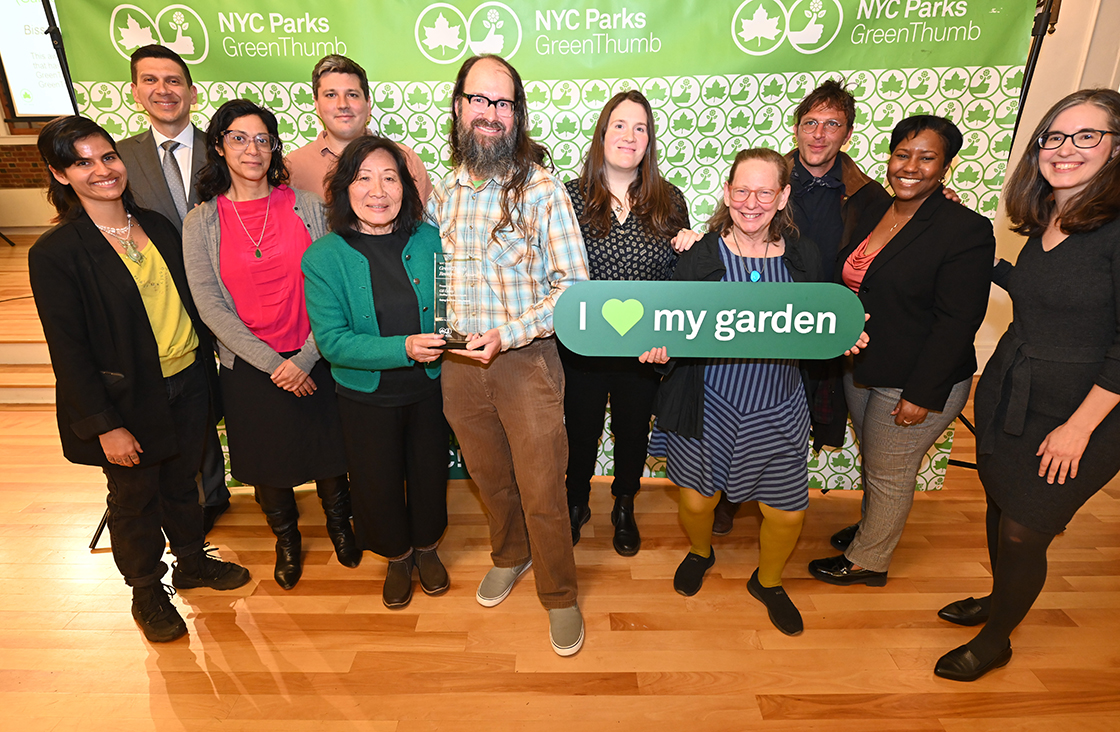 NYC Parks staff and honorees of the GreenThumb Going the Extra Mile Award are standing in front of a bright green and white NYC Parks GreenThumb step and repeat at the 2023 Garden Recognition Awards ceremony. A person is holding the glass award and one person is holding a sign saying I heart my garden.