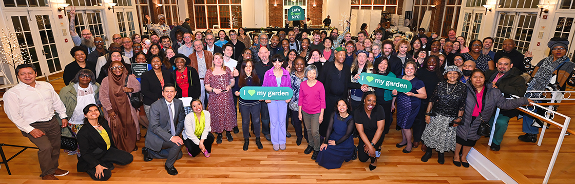 A large group of smiling people standing, sitting, and kneeling in a large bright room. Some people are holding garden recognition awards and signs saying I heart my garden.