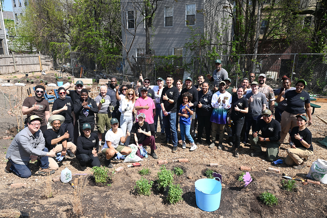 A large group of smiling people standing and kneeling in rows in a garden with their thumbs up. In front of the group of people are plants on the ground and garden tools.