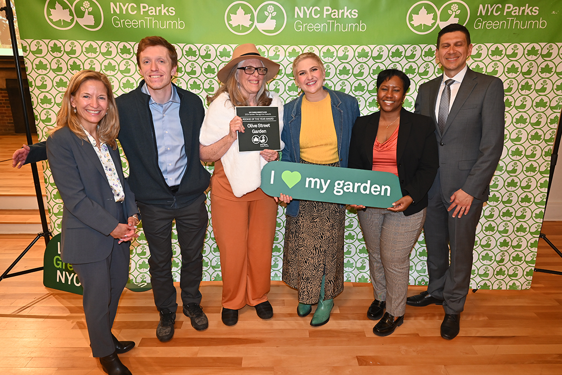 NYC Parks staff and honorees of the GreenThumb Rookie of the Year Award are standing in front of a bright green and white NYC Parks GreenThumb step and repeat at the 2023 Garden Recognition Awards ceremony. A person in the center is holding the award, a hunter green plaque, and one person is holding a sign saying I heart my garden.