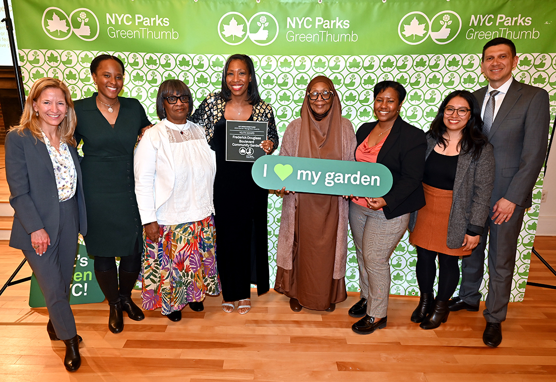 NYC Parks staff and honorees of the GreenThumb Youth Award are standing in front of a bright green and white NYC Parks GreenThumb step and repeat at the 2023 Garden Recognition Awards ceremony. A person in the center is holding the award, a hunter green plaque, and one person is holding a sign saying I heart my garden.