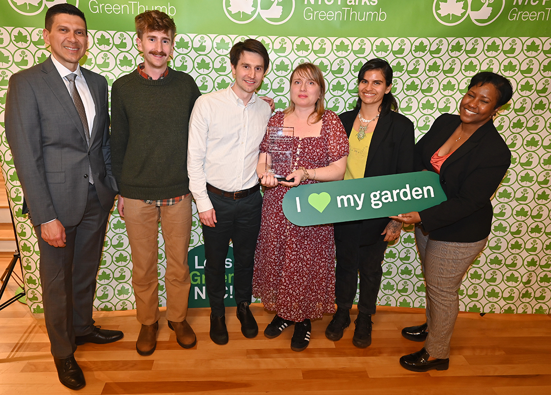 NYC Parks staff and honorees of the GreenThumb Going the Extra Mile Award are standing in front of a bright green and white NYC Parks GreenThumb step and repeat at the 2023 Garden Recognition Awards ceremony. A person in the center is holding the glass award and one person is holding a sign saying I heart my garden.