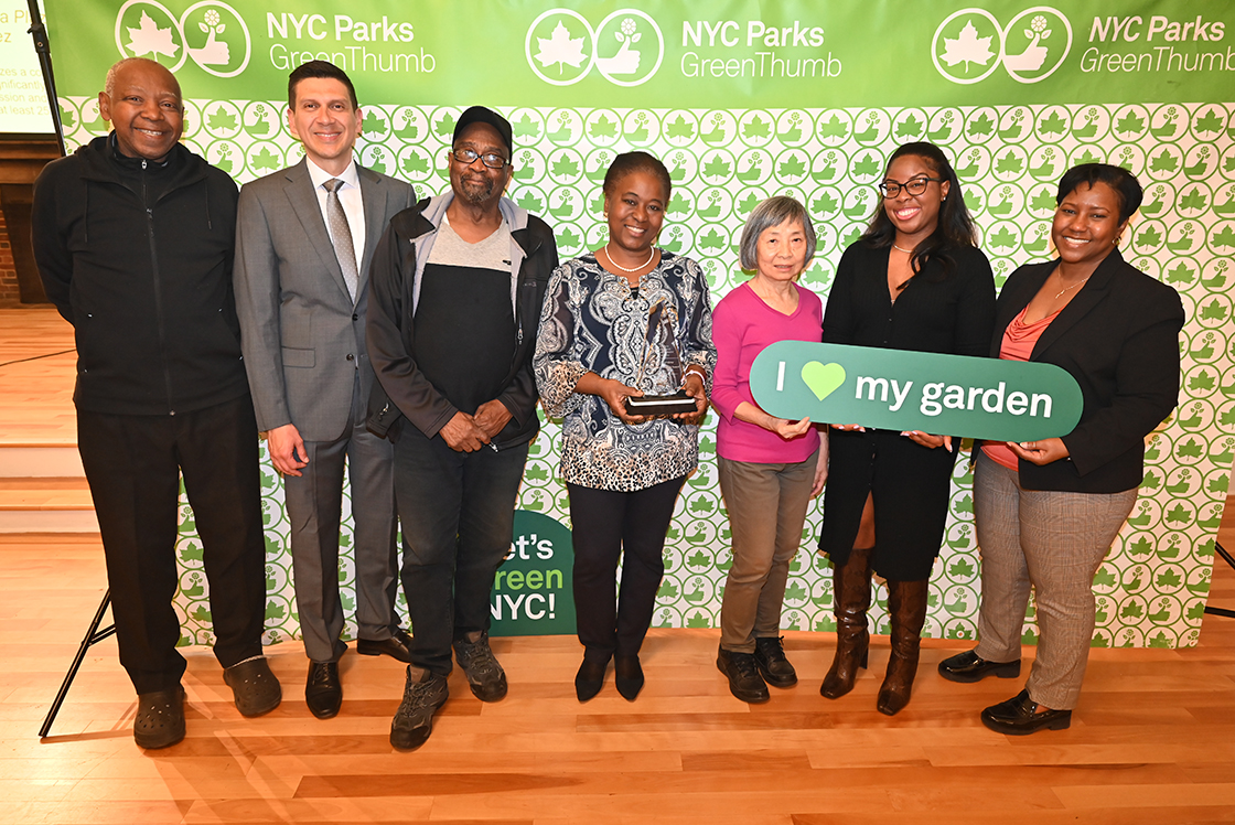 NYC Parks staff and honorees of the GreenThumb Garden Group Lifetime Achievement Award are standing in front of a bright green and white NYC Parks GreenThumb step and repeat at the 2023 Garden Recognition Awards ceremony. A person is holding the glass award and one person is holding a sign saying I heart my garden.