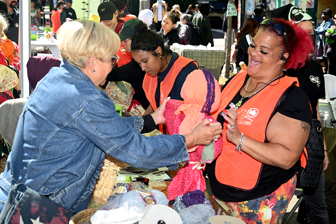 Two women are facing each other at a table. The woman on the left side holds a fabric scarecrow as the woman on the right side helps her decorate it with a smile.