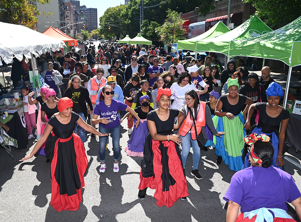 A group of people dancing in the middle of a block during a street fair with tables and tents on each side.