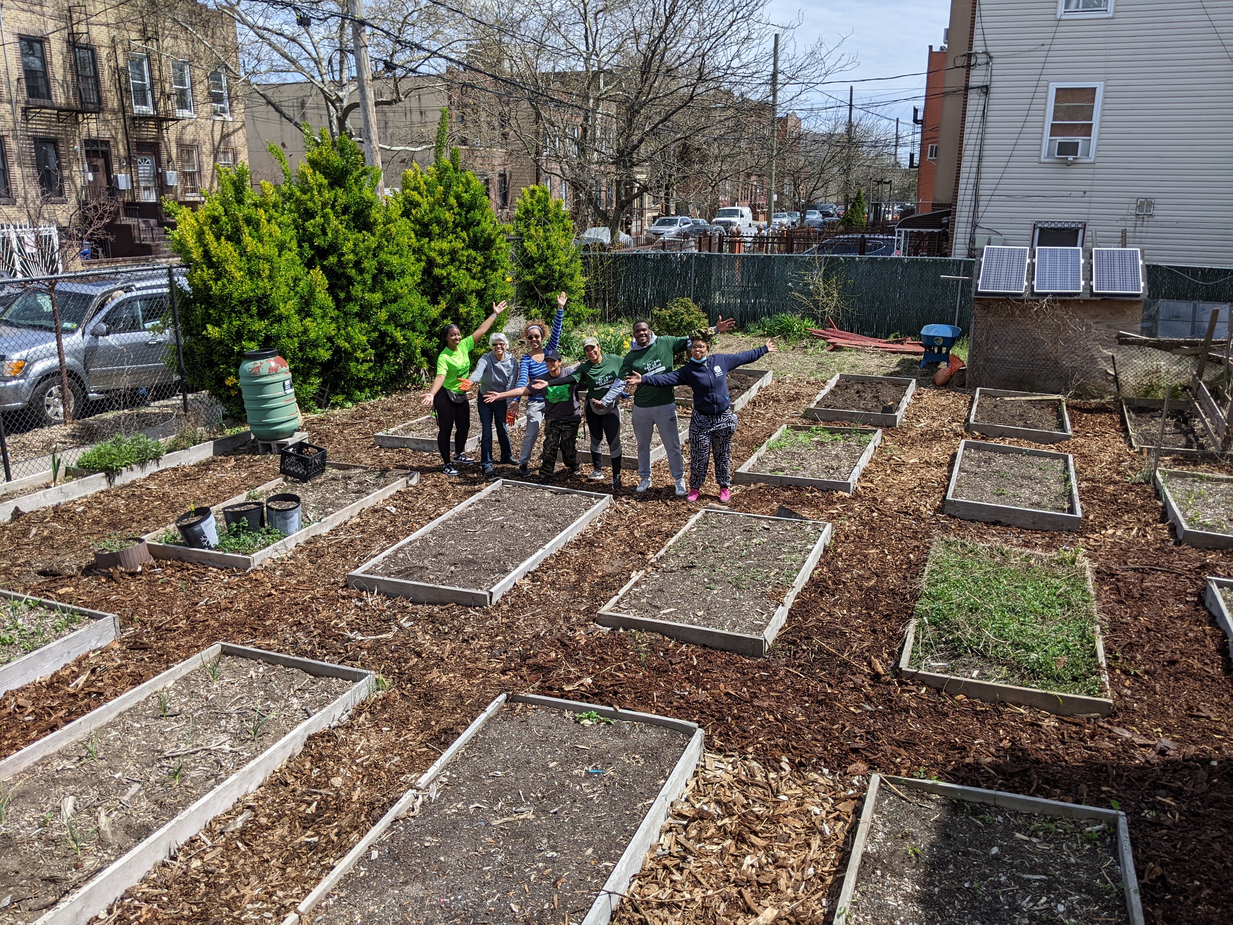 A community garden with rows of wood raised garden beds that are filled with soil, seven people are standing in a line in the middle of the garden. Each person has one arm out in front of them and the other raised high in unison.