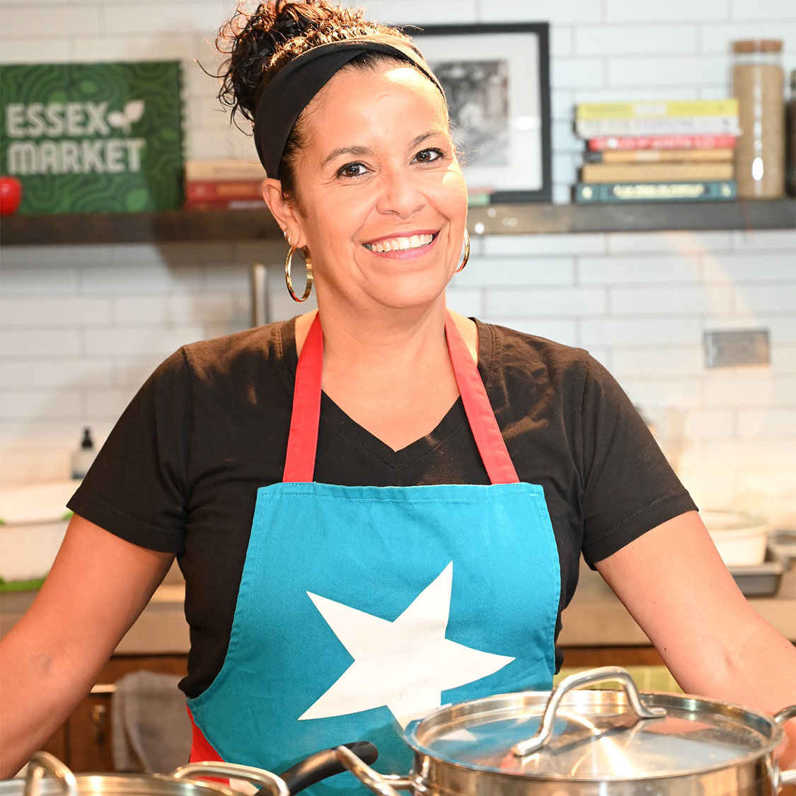 A woman with dark brown hair in a ponytail smiles at the camera. She is standing in front of cooking pots and wearing a black t-shirt and an apron with the flag of Puerto Rico.