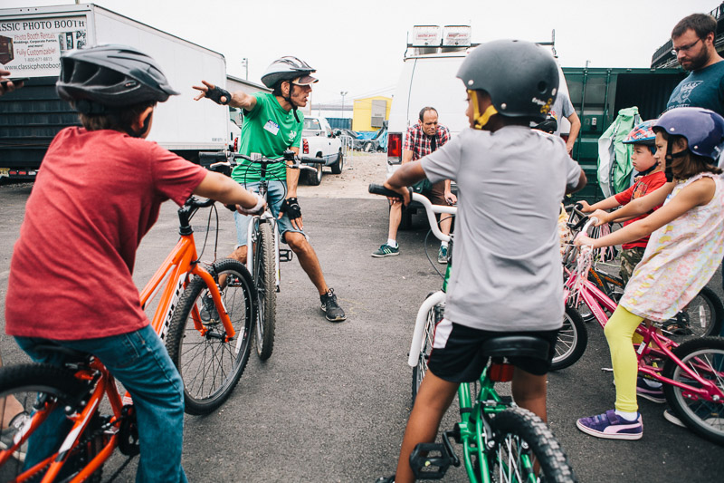 A group of young kids on bicycles with their backs to the camera as they listen to an instructor teaching them bike skills.